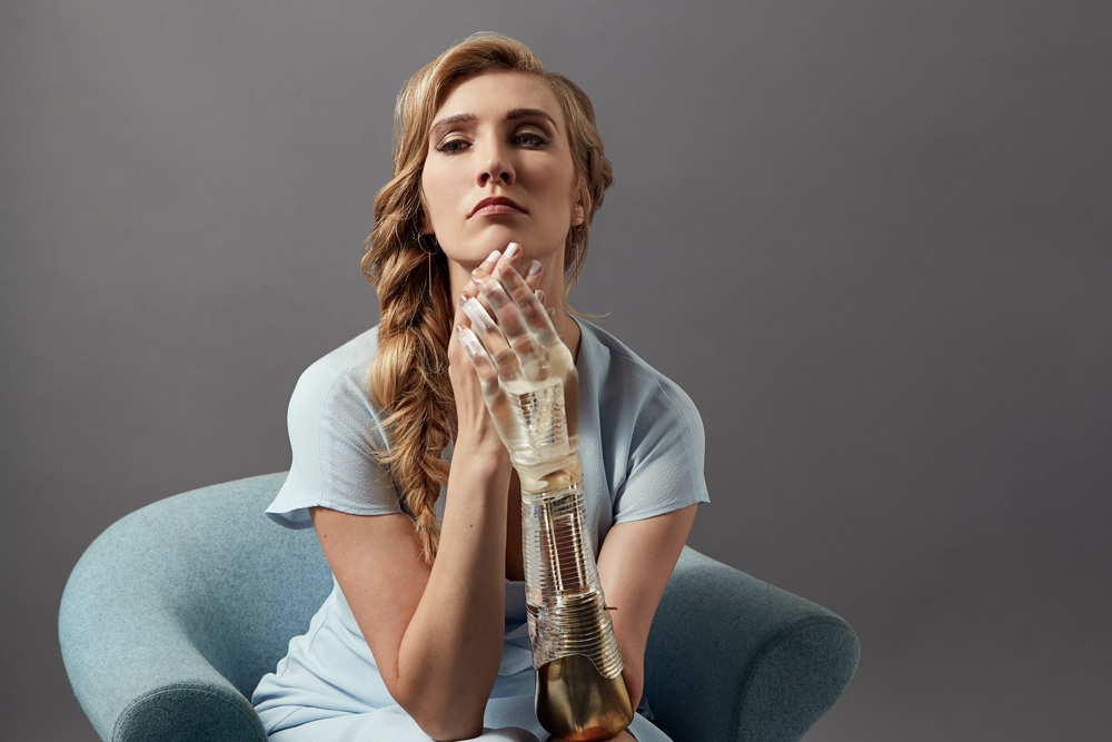 A woman crossing arms in front of her face. One arm is a transparent and gold prosthetic arm.